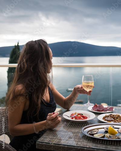 Young woman sits on the balcony at the table with oysters, meat and salad and holds glass with sparkling wine. Elegant girl is looking on the calm sea and mountains on the background. Side view