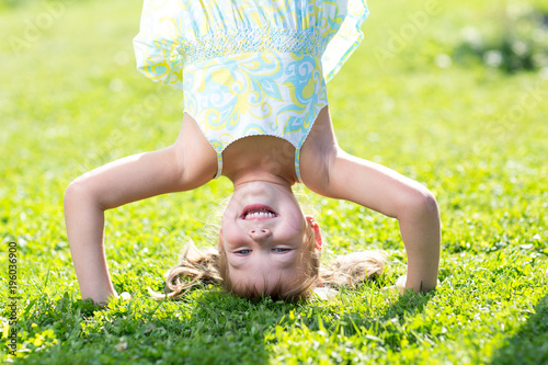 Happy little kid girl standing on her head on green lawn.