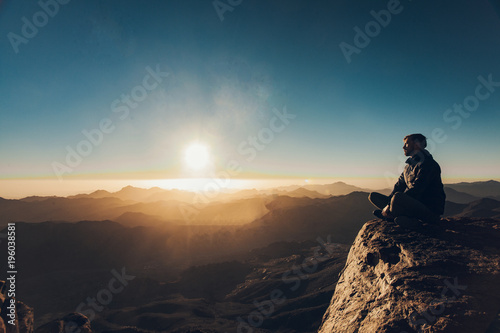 Man sits in a pose of yoga on Mount Sinai and meditates against background of sunrise.