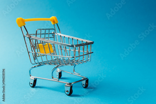 Close up of supermarket grocery push cart for shopping with black wheels and yellow plastic elements on handle isolated on blue background. Concept of shopping. Copy space for advertisement