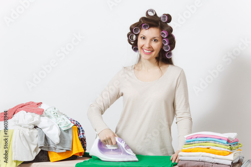 Young pretty housewife with curlers on hair in light clothes ironing family clothing on ironing board with iron. Woman isolated on white background. Housekeeping concept. Copy space for advertisement.