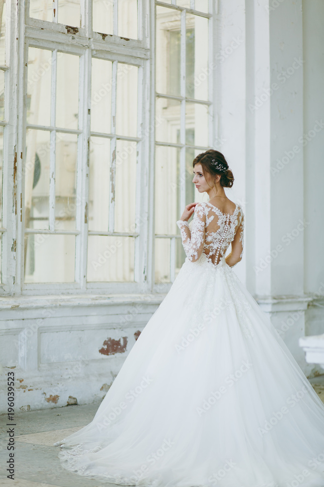 Beautiful wedding photosession. Young bride in a white lace dress with a long plume with an exquisite hairstyle in vintage interior on the veranda of an old house with columns near the garden