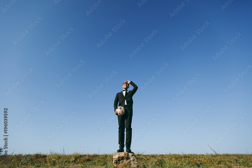 Beautiful wedding photosession. Handsome bridegroom in a black suit and white shirt with a bouquet of the bride stands on a stone on walk around the big green field against blue sky background Beautiful wedding photosession. Handsome bridegroom in a black suit and white shirt with a bouquet of the bride stands on a stone on walk around the big green field against blue sky background