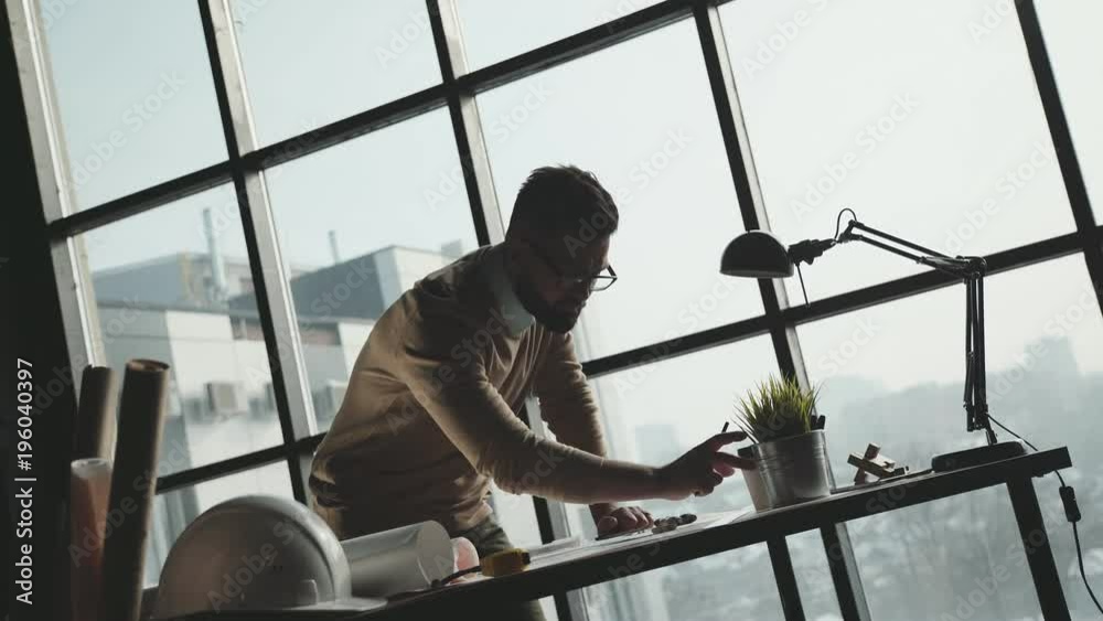 Engineer works in a bright office with a large window, concentrates and draws blueprints. Workplace of an architect or designer: loft style, minimalistic interior, drawings and coffee on the table. Engineer works in a bright office with a large window, concentrates and draws blueprints. Workplace of an architect or designer: loft style, minimalistic interior, drawings and coffee on the table.