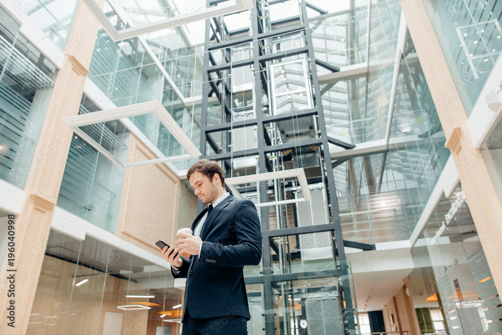 Coffee break on go. confident man holding coffee cup and using his smart phone while walking in hall