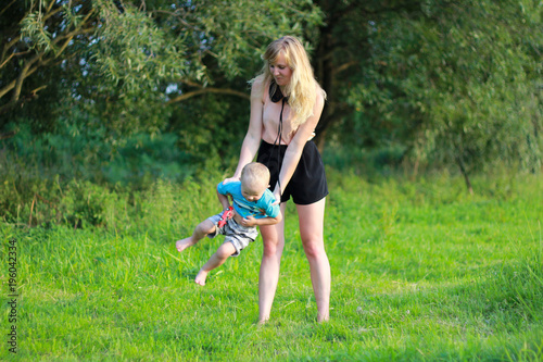Young blonde mother in playing with her child in the green lawn. Happy fun summer photo. Smiling laughing boy with his mom. Sunny day outdoors in nature.