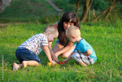 Mother with dark hair is playing with two children in the green lawn. Happy fun summer photo. Smiling laughing boys with his mom. Sunny day outdoors in nature. Kids and mom are hugging.