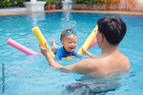 Cute little Asian 18 months / 1 year old toddler boy child wear swimming goggles learning to swim with pool noodle at outdoor pool; dad and son relaxing in swimming pool of clubhouse in summer day