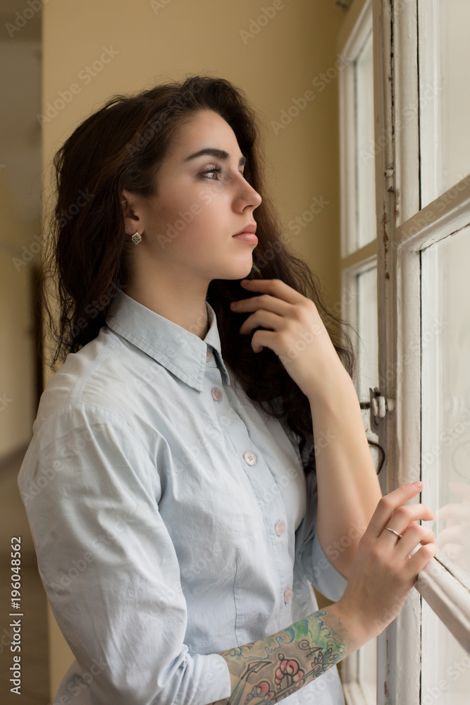 Fabulous young woman with curly hair and a tattoo on her arm posing near the window Fabulous young woman with curly hair and a tattoo on her arm posing near the window