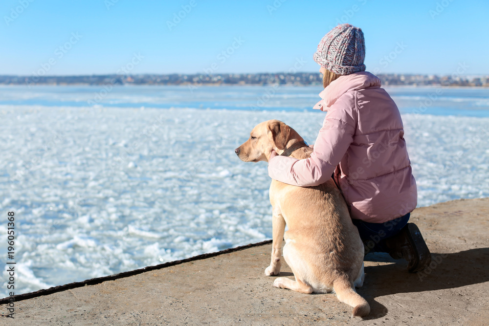 Woman with cute dog near river on winter day. Friendship between pet and owner