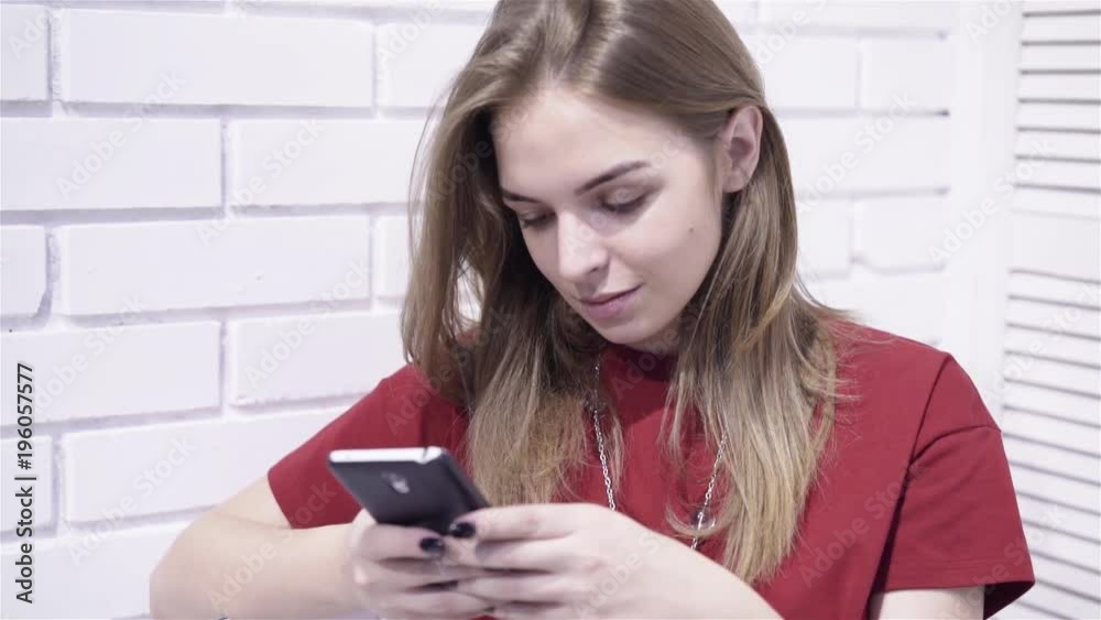 Calm young woman wearing a red t shirt and trousers is sitting on a chair in a room and texting. Handheld slow motion zoom in shot Calm young woman wearing a red t shirt and trousers is sitting on a chair in a room and texting. Handheld slow motion zoom in shot
