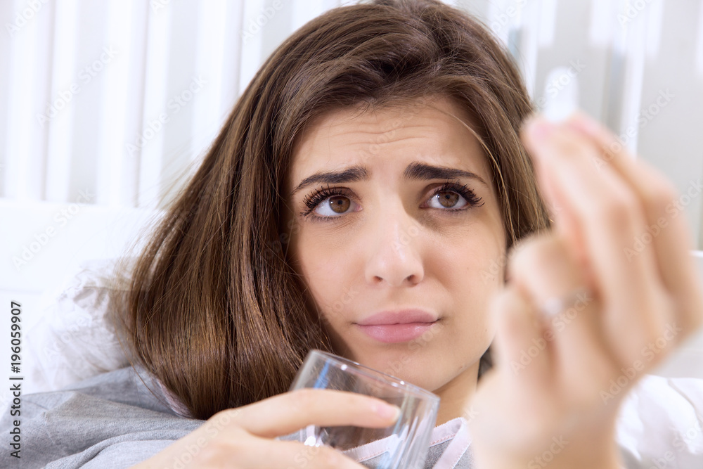 Sick young woman in bed looking pill medicine funny expression Sick young woman in bed looking pill medicine funny expression
