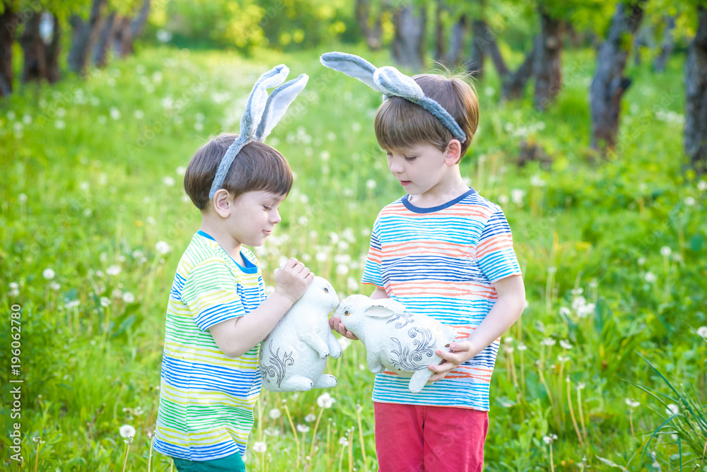 Two little kids boys and friends in Easter bunny ears during traditional egg hunt in spring garden, outdoors. Siblings having fun with finding colorful eggs. Old christian catholoc tradition