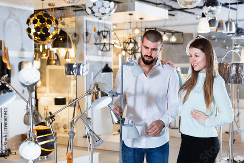 Smiling couple customers choosing floor lamp with a long standard in store