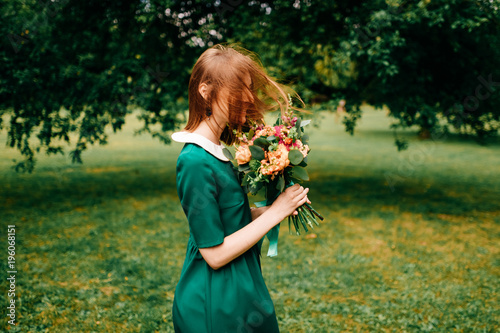 Young happy red haired girl with bouquet of summer flowers enjoying inner peace in green dress at nature. Little child playing in wonderland park. Pretty woman portrait outdoor. Lovely smiling face.