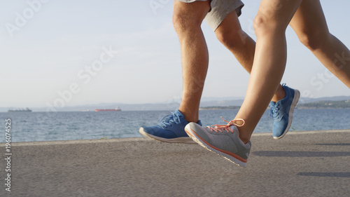 LOW ANGLE: Unrecognizable sporty couple jog along the beautiful sunny coastline.