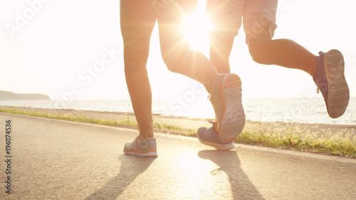 Fototapeta Naklejka Na Ścianę i Meble -  LOW ANGLE, SUN FLARE: Young athletic woman and man run along sunny ocean road.