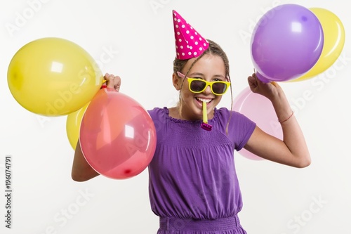 Cheerful teenage girl 12,13 years old, with balloons, in festive hat, blowing a pipe on white background,