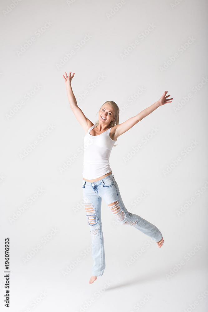 Fototapeta premium Young smiling woman in jeans and a jersey jumping against a gray background.