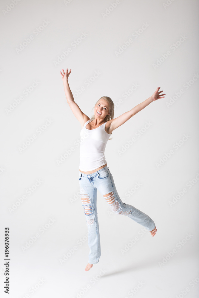 Young smiling woman in jeans and a jersey jumping against a gray background.