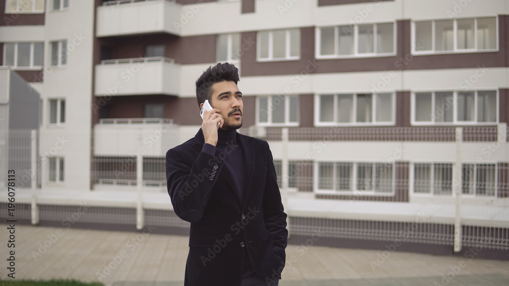Portrait of attractive slender businessman in formal suit talking on smartphone against the backdrop of business center