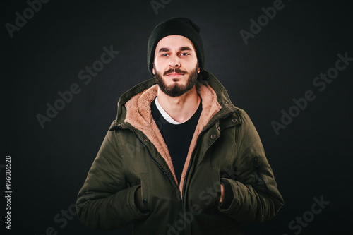 Young bearded man in denim jacket with shearling liner shows his chest wearing black unlabeled henley shirt longsleeve, isolated on grey wall in studio looks into camera