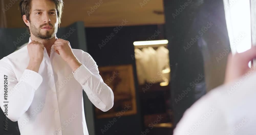 A handsome man in his loft dresses his sexy white shirt in a sexy way, looks into his room and smiles with a sure look at himself. Concept of: elegance, business, work.