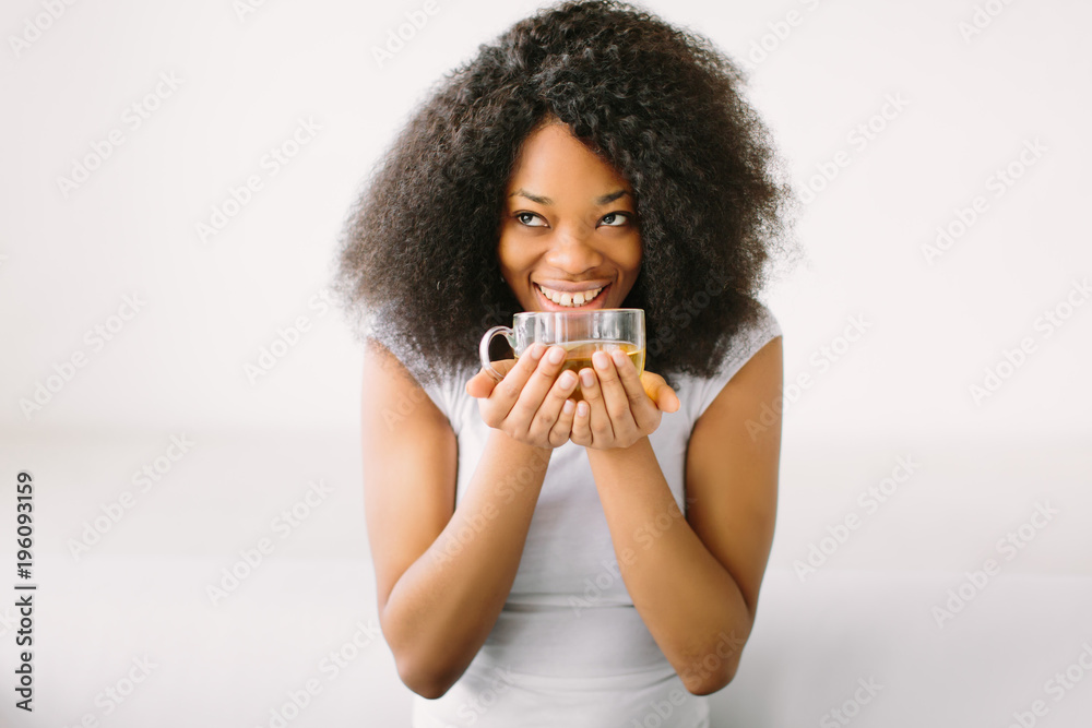 An African American young woman sitting in the lotus position on white bed with cup of tea An African American young woman sitting in the lotus position on white bed with cup of tea