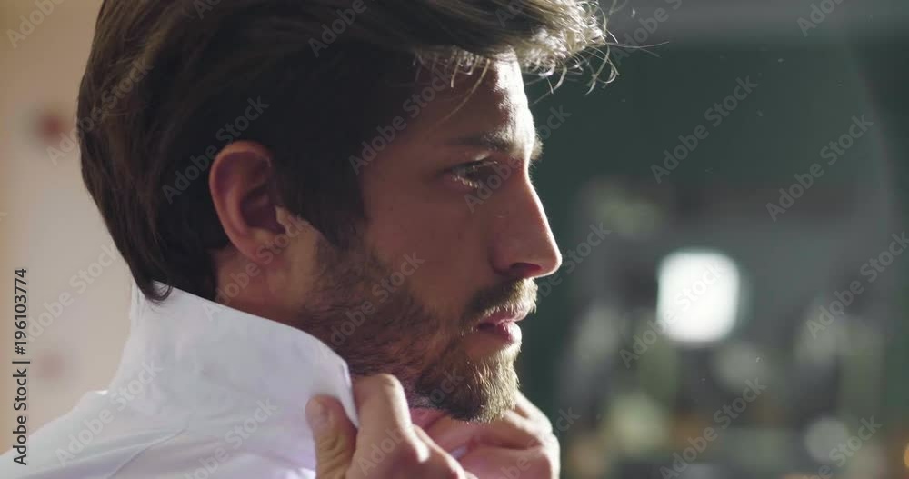 A handsome man in his loft dresses his sexy white shirt in a sexy way, looks into his room and smiles with a sure look at himself. Concept of: elegance, business, work.