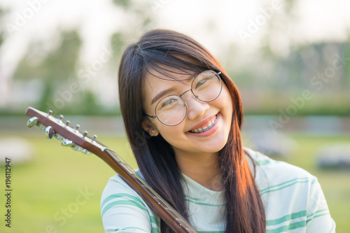 Asian teen girls  with a guitar at the shoulder in the lawn. She is wearing braces  and wear eyeglasses.
