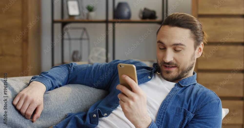 Portrait of the attractive man in jeans shirt resting on the sofa and texting and taping on the smartphone in the living room. Indoors Portrait of the attractive man in jeans shirt resting on the sofa and texting and taping on the smartphone in the living room. Indoors