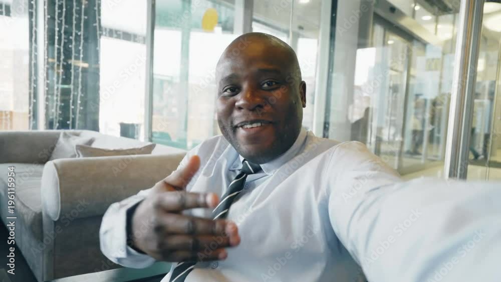 Point of view shot of businessman in formal wear having online video chat with family on smartphone and waving his hand happily in a classy cafe. He smiles cheerfully.