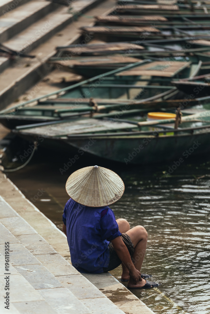 Back of woman wearing purple shirt, conical hat carry wash her feet in the river with empty rowing boats in the background at Trang An Grottoes in Ninh Binh, Vietnam.