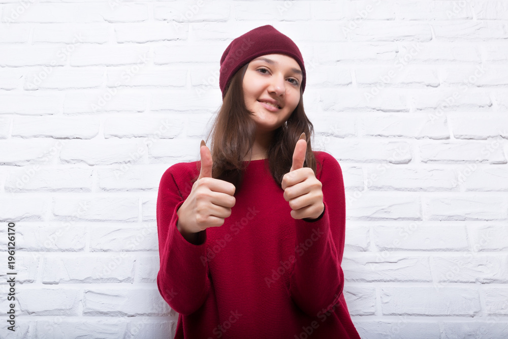 A young pretty smiling girl in a red sweater and a hat against a modern white brick wall in the loft style. She looks at the viewer. Copy space. A young pretty smiling girl in a red sweater and a hat against a modern white brick wall in the loft style. She looks at the viewer. Copy space.