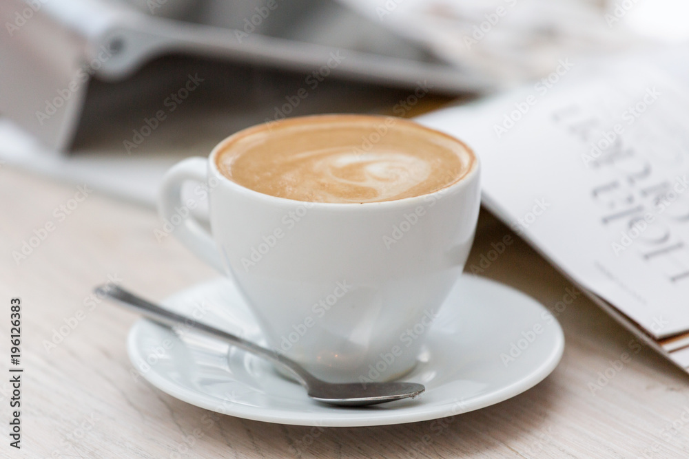 A white cup with a coffee drink, standing next to a tablet and magazine. Bright interior.