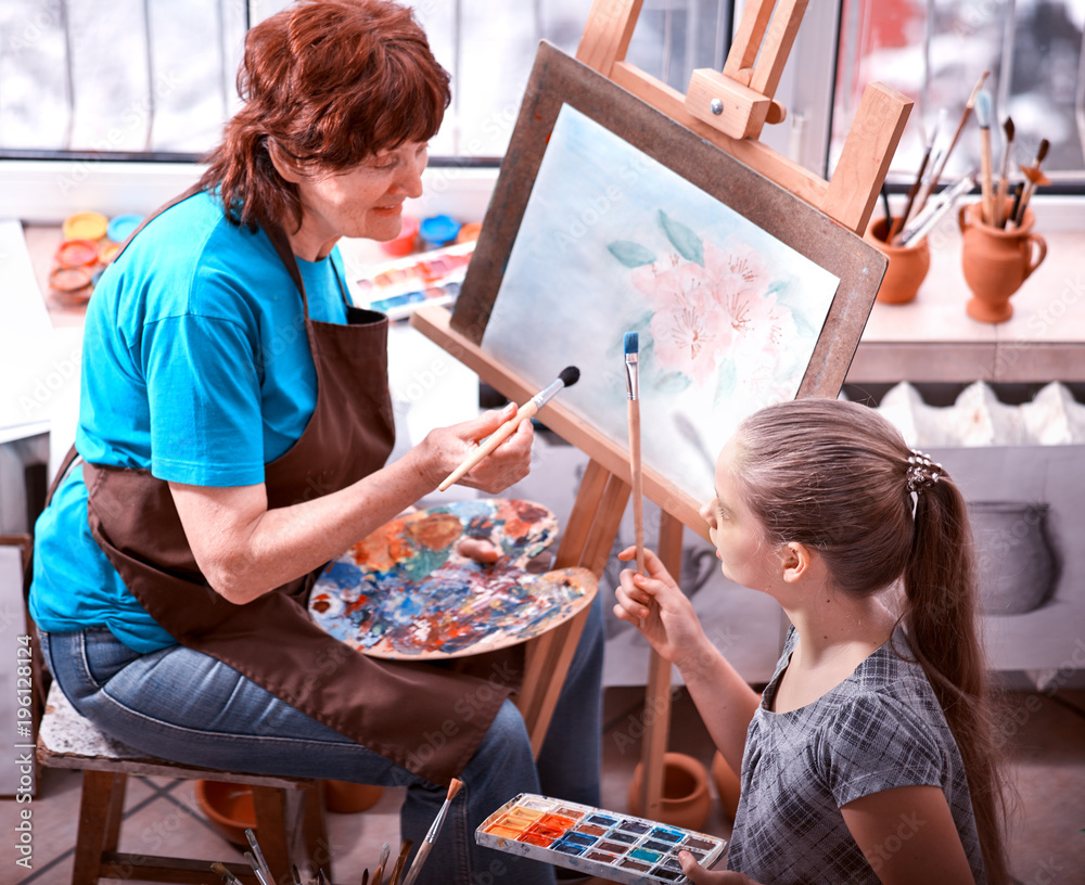 Obraz premium Artist painting easel in studio. Authentic grandmother and kids girl paints with palette watercolor paints palette and brush morning sunlight. Courses on drawing for children and adults.