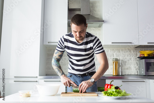 Young man preparing delicious and healthy food with vegetables in the home white kitchen