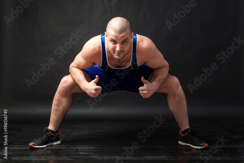 A young athletic man in blue wrestling tights and blue shorts makes a squat with a wide leg arrangement and shows thumbs up on a black isolated background in a photo studio