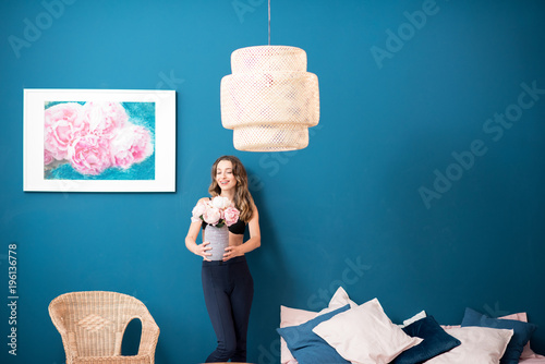 Beautiful young woman holding flowers standing in the cozy bedroom on the blue wall background at home
