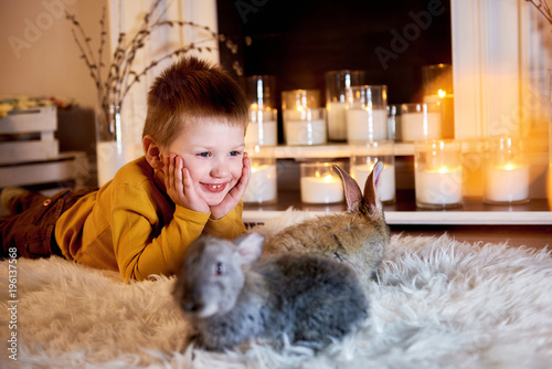 Little boy with rabbit. Blond nice boy lying happily with grey rabbits around and warm candles behind him