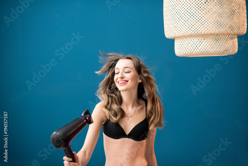 Beautiful woman in bra drying hair with hairdryer standing on the blue wall background at home