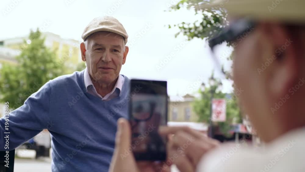 Senior man taking smartphone picture of his friend, who is sitting on bench in pedestrian street Senior man taking smartphone picture of his friend, who is sitting on bench in pedestrian street