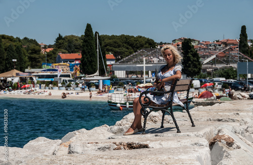 A young european blonde woman sits on the bench near the Mediterranean Sea.