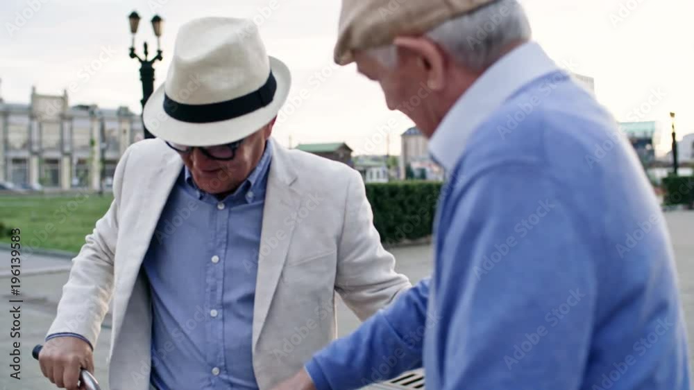 Two senior male friends shaking hands when meeting each other in the city, one of them showing his bicycle to the other one Two senior male friends shaking hands when meeting each other in the city, one of them showing his bicycle to the other one