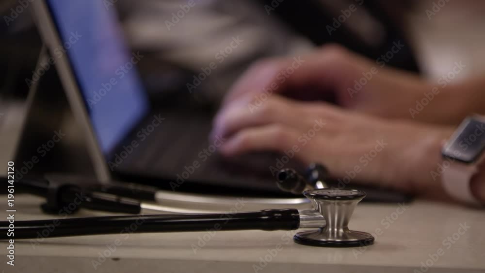 View of stethoscope on desktop as hands are typing in backgound on laptop in medical clinic. View of stethoscope on desktop as hands are typing in backgound on laptop in medical clinic.