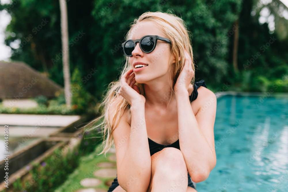 Stylish young woman with long blond hair dressed in swimsuit and sunglasses has fun by the pool. Modern enjoyable caucasian woman relaxes outdoors on the background of the jangles Stylish young woman with long blond hair dressed in swimsuit and sunglasses has fun by the pool. Modern enjoyable caucasian woman relaxes outdoors on the background of the jangles