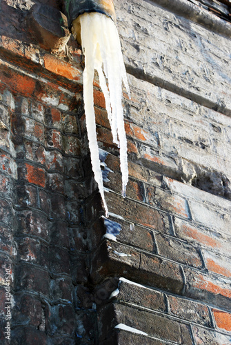 Icicle from drain pipe, wall with red bricks under ice, grunge background, vertical