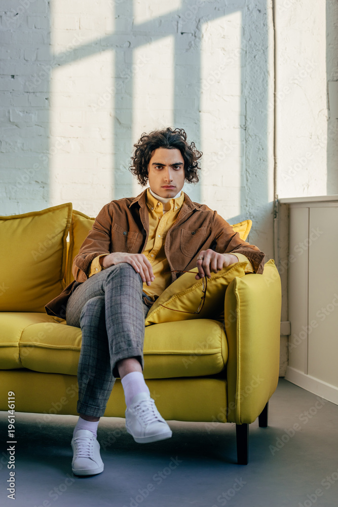 Young stylish man with eyeglasses in hand sitting on couch