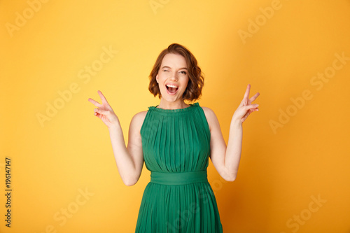 portrait of young happy winking woman showing peace sign isolated on orange