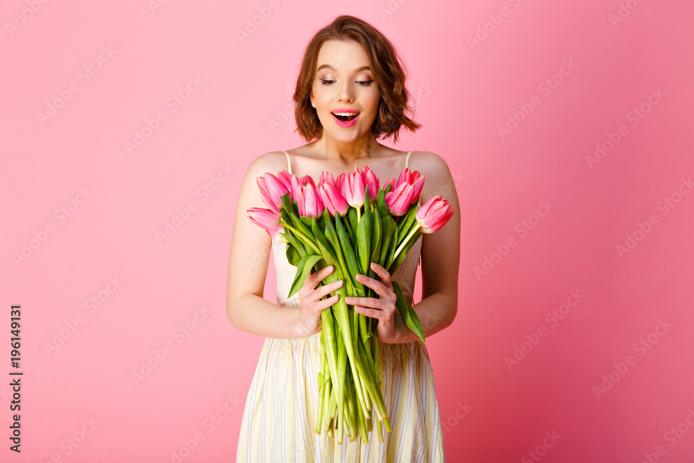 portrait of surprised woman looking at bouquet of pink tulips in hands isolated on pink